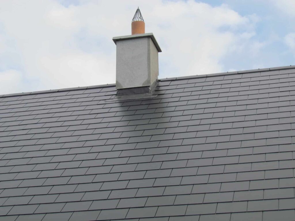Front elevation of a completed grey slate pitched roof on a rural residential property in Ireland, showing uniformly laid slate tiles across the full width, a rendered chimney stack with a terracotta flue liner and decorative wire bird guard, and lead stepped flashing at the chimney base against a partly cloudy sky.