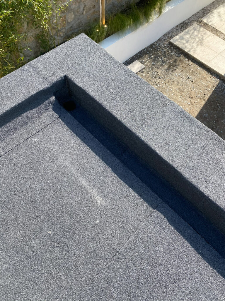 Close-up corner detail of a completed dark grey mineral-finish felt flat roof in Ireland, showing a precisely formed internal corner upstand with a recessed drainage channel, and a landscaped garden with stone wall and ornamental grasses visible below.