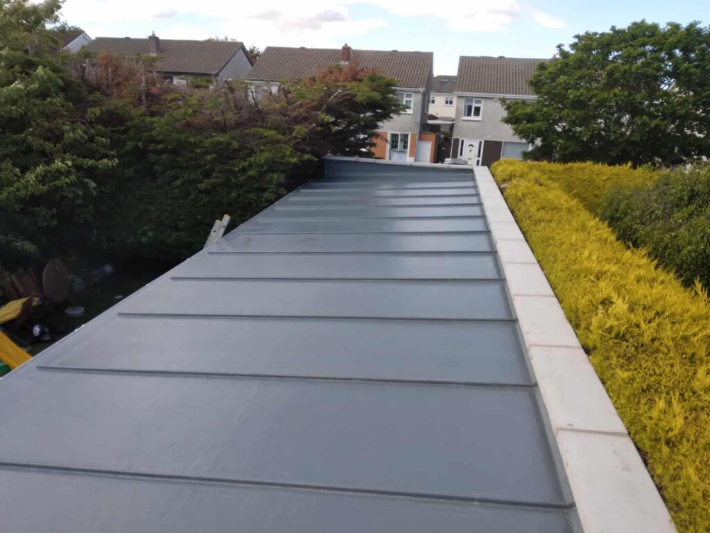 Aerial view along the length of a completed standing seam metal roof in dark grey-blue, showing parallel raised seam profiles running the full slope from ridge to eaves. White stone coping is visible along the right-hand verge. Residential properties and garden hedging are visible in the background.