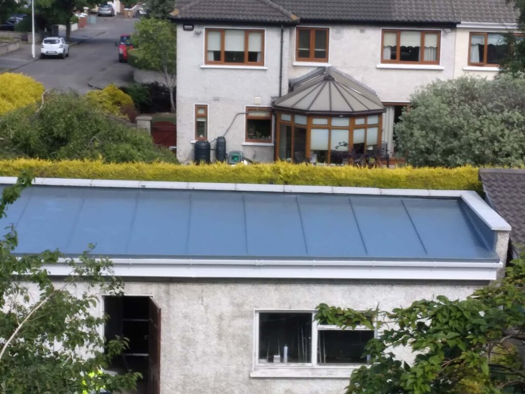 Elevated external view of a single-storey residential extension with a newly completed standing seam metal roof in dark grey-blue. The full roof slope is visible from above, showing evenly spaced raised seams running from ridge to eaves and a white aluminium or UPVC fascia and gutter system at eaves level. The host dwelling is rendered in white with timber-framed windows. Neighbouring properties, mature hedging, and a residential street are visible in the background.
