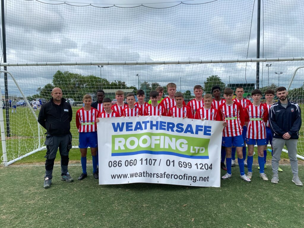 A youth football team of approximately fifteen players in red and white striped kits standing in front of a goal net, holding a Weathersafe Roofing Ltd banner displaying the company's phone numbers and website address, with two adults in non-playing attire flanking the group on an outdoor pitch in Ireland.