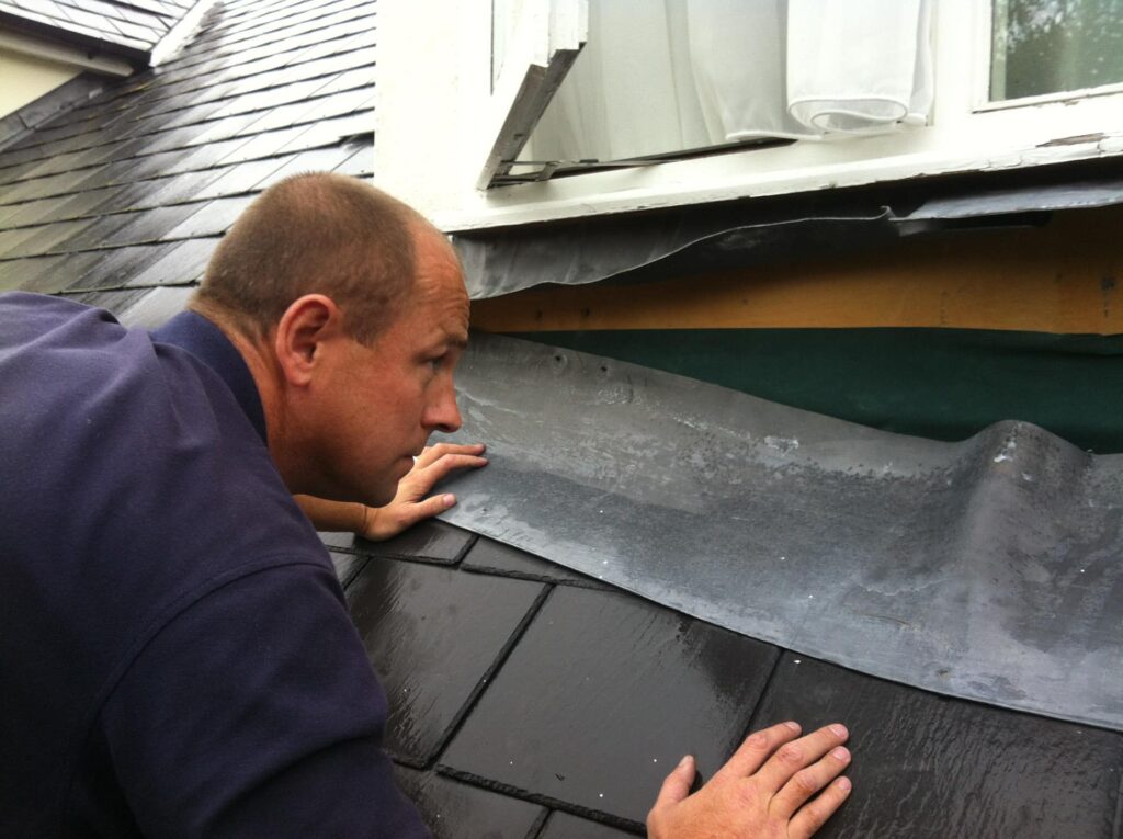 A Weathersafe Roofing tradesperson kneeling on a dark grey natural slate roof in Ireland and closely inspecting a failed lead flashing at the abutment between the roof and a dormer window frame, with lifting and cracked lead visible at the junction and an existing slate roof on the adjoining property visible behind.