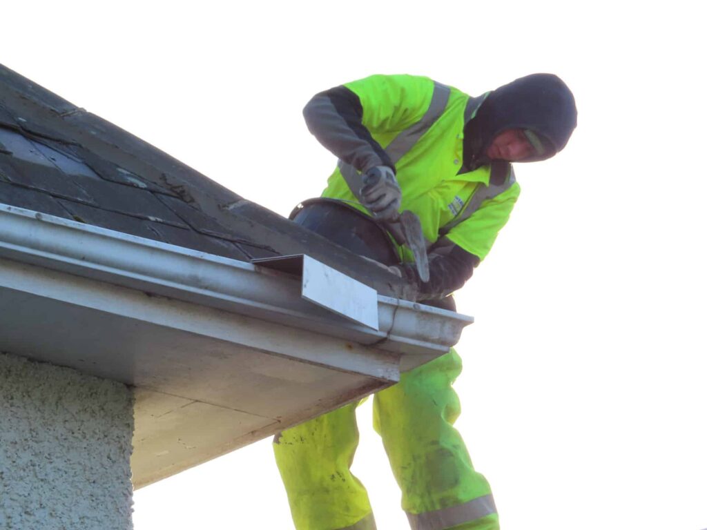 A Weathersafe Roofing tradesperson in a bright yellow high-visibility jacket and hat working at the eaves of a residential slate roof in Ireland during cold weather, using a hammer to fix a lead or zinc gutter bracket to a grey aluminium fascia gutter, with a rendered gable wall visible below.