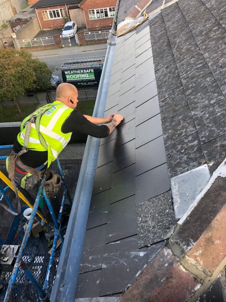 A Weathersafe Roofing tradesperson in a high-visibility vest standing on scaffolding and reaching over a zinc or lead box gutter to fix dark grey fibre cement slates to the eaves course of a residential roof in Ireland, with an existing weathered natural slate roof to the right and the Weathersafe Roofing Ltd branded company van visible parked on the street below.