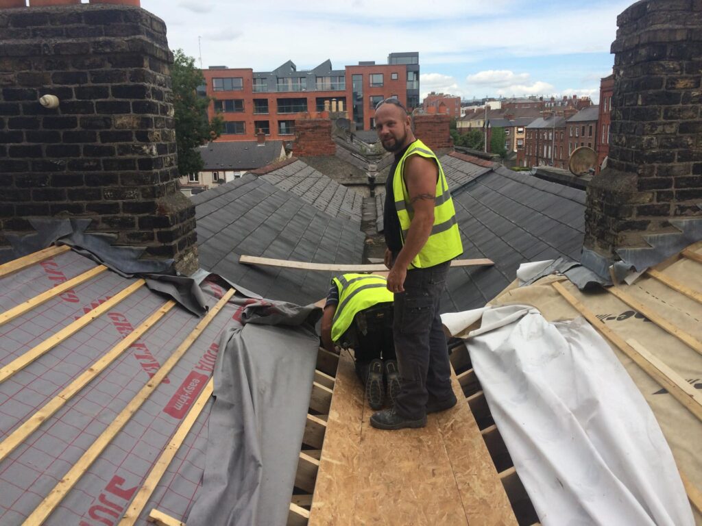 Two Weathersafe Roofing tradespeople in high-visibility vests working on a complex multi-pitch slate roof at a valley junction on an urban residential property in Ireland, with new breathable underlay, OSB decking, and timber battens visible, and Dublin city rooftops in the background.