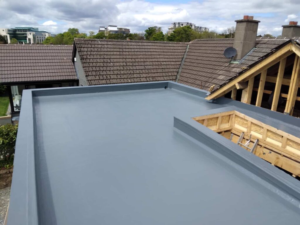 Roof level view of a completed GRP fibreglass flat roof in grey showing a rectangular rooflight upstand curb to the right, fibreglass-coated and awaiting glazing. The roof deck surface is smooth and seamless with fully formed upstands along all parapet edges. New timber roof structure is visible at the upper right. Neighbouring tiled rooftops and a commercial building are visible in the background.