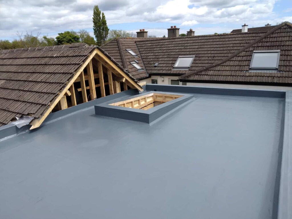 Aerial view of a completed GRP fibreglass flat roof in grey, showing a fully formed square rooflight upstand curb in the centre of the roof deck. The upstand is fibreglass-coated with the timber rooflight frame exposed and awaiting glazing. New timber roof structure forming a pitched gable is visible to the left. Neighbouring pitched tile roofs with roof windows are visible in the background.