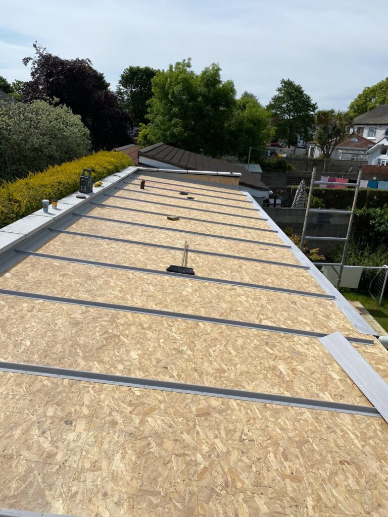 : Aerial view of a pitched roof slope fully covered with OSB oriented strand board decking, laid in staggered coursed panels. Evenly spaced metal z-section or counter batten profiles are visible running down the slope between the board panels. A broom, work radio, and paint tin are visible on the surface. A ladder and scaffold frame are visible at the right-hand eaves edge.