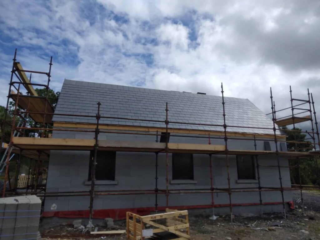 External view of a single-storey new-build residential property with a completed natural slate roof, showing the full front elevation. The slate roof slope is fully laid in a coursed staggered bond pattern in blue-grey. The blockwork walls are rendered in grey. Window and door openings are formed but not yet glazed or fitted. Multi-level scaffold remains in place on both gable ends. Timber fascia boarding is visible at the eaves.