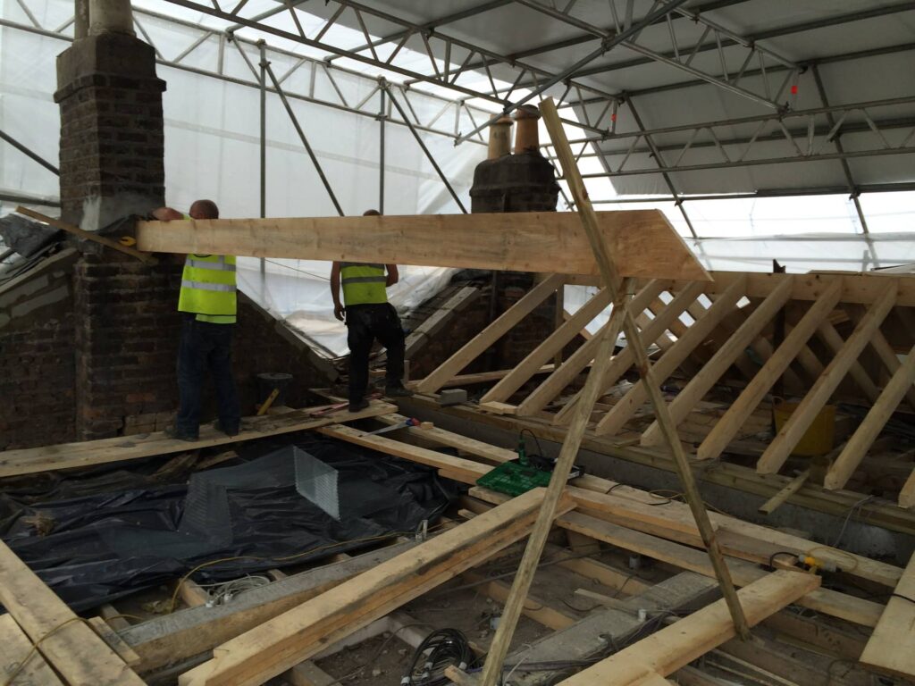 Two Weathersafe Roofing tradespeople in high-visibility vests lifting and positioning a large timber ridge beam inside a temporary weather enclosure over a residential property in Ireland, with new cut roof framing, brick chimney stacks with terracotta pots, polythene enclosure panels, and temporary protection sheeting all visible.