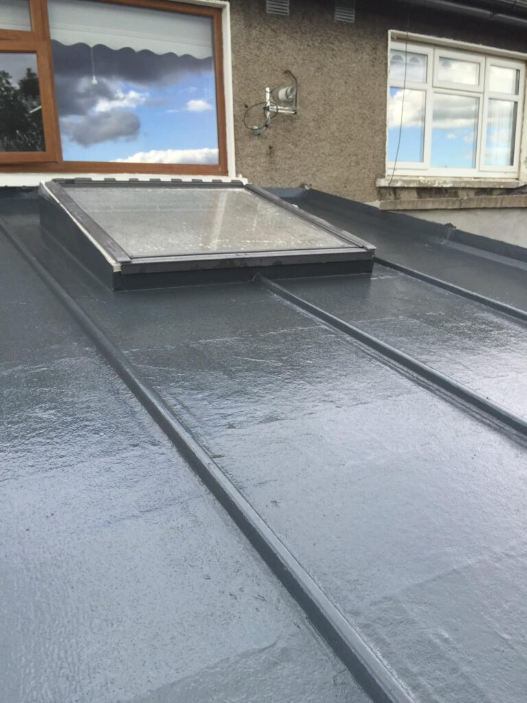 Completed dark grey GRP fibreglass flat roof on a residential extension in Ireland, showing raised drainage channels running the length of the deck, a low-profile framed rooflight set flush into the surface, and a rendered gable wall with timber-framed and uPVC windows above.