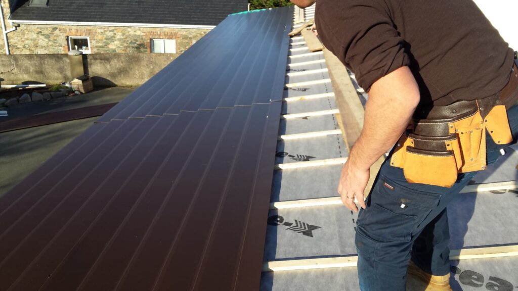 A roofer wearing a tool belt fitting dark brown profiled metal roof cladding panels to a residential roof in Ireland, with breathable underlay and evenly spaced timber battens visible on the unclad section, and a stone-built neighbouring property in the background.