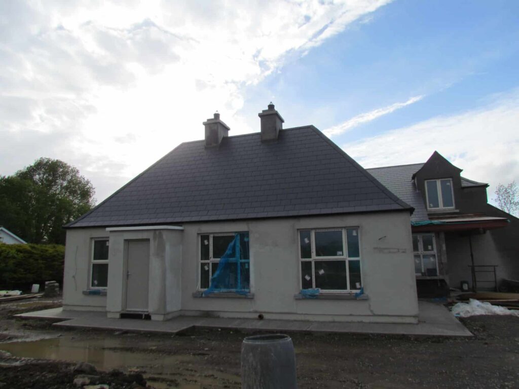 Front elevation of a newly rendered single-storey bungalow in Ireland with a fully completed dark grey slate hip roof, twin rendered chimney stacks with bird guards, new uPVC windows still carrying protective film, and a two-storey dormer extension visible to the right under construction.