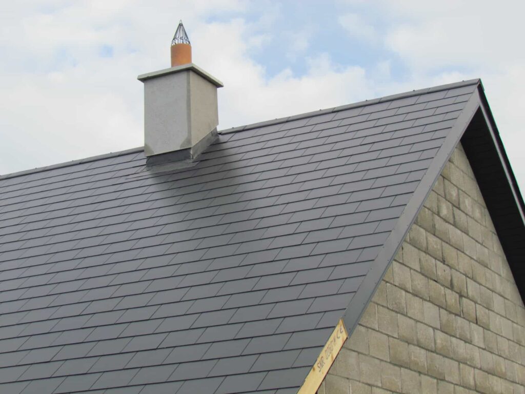 Close-up of a completed dark grey slate gable end roof on a rural new-build property in Ireland, showing uniformly laid slates, a black uPVC verge trim, lead flashing at the base of a rendered chimney stack with a terracotta flue liner and decorative wire bird guard, against a partly cloudy sky.
