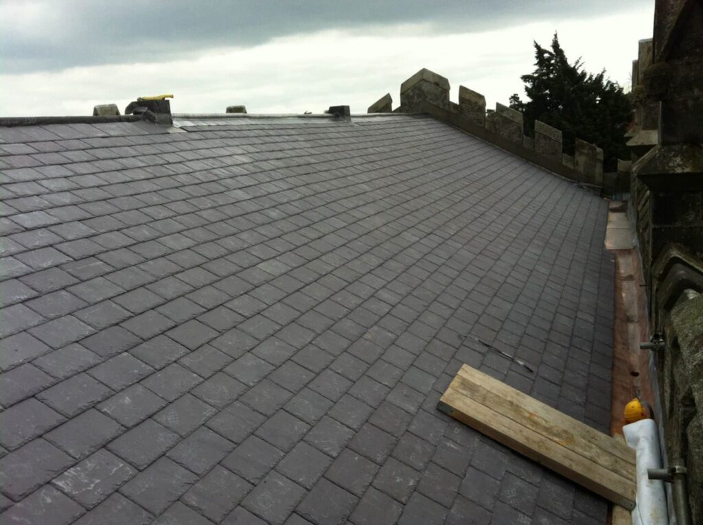 Aerial view of a large completed grey natural slate roof on a historic church or institutional building in Ireland, showing uniformly laid slates across the full pitch with a lead parapet gutter along the right-hand edge, a stone battlemented parapet wall, and overcast sky, with timber planks and materials staged at the eaves indicating works nearing completion.