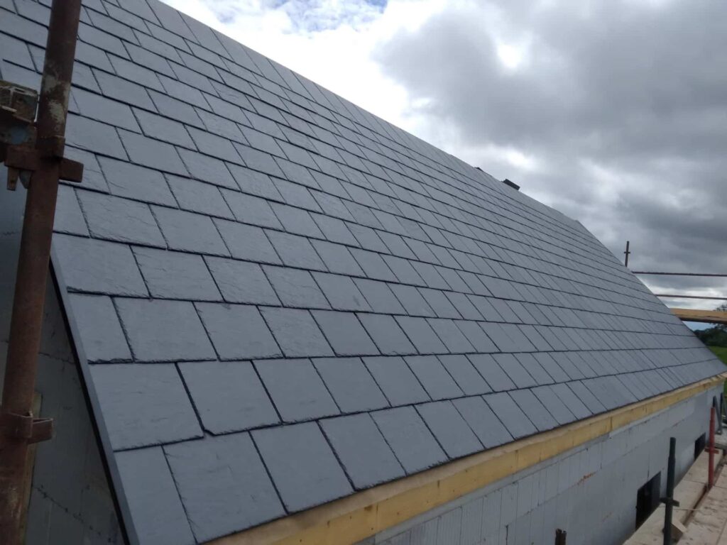 Roof level view of a large completed natural slate roof slope in blue-grey, showing the full extent of coursed staggered bond slating from eaves to ridge. The slates are uniformly laid with consistent lap and bond. A small lead or GRP pipe penetration flashing is visible near the ridge. Timber fascia and insulation are visible at the eaves. Scaffold is visible to the upper left. An overcast sky is visible above.