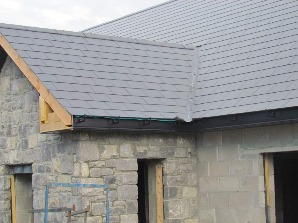Side elevation of a new-build rural property in Ireland with a completed grey slate roof showing a lead-dressed external valley, black uPVC fascia and gutter system fitted to the eaves, a timber verge detail to the gable, and an unrendered limestone and concrete block wall with open window and door openings below.