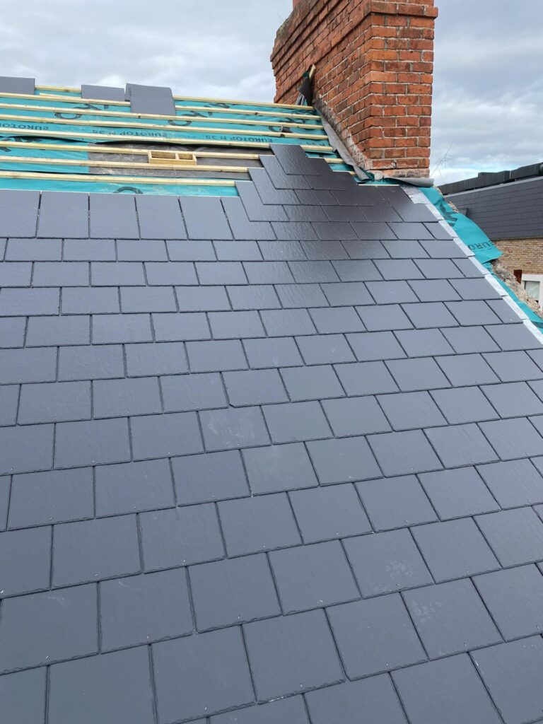 Close-up view of a pitched roof slope in the process of being re-slated with new dark grey fibre cement or artificial slates in a staggered coursed bond pattern. The upper portion of the slope shows Eurotop N35 breathable felt and counter battens with slating in progress. A red brick chimney stack is visible at the upper right. An overcast sky is visible above.