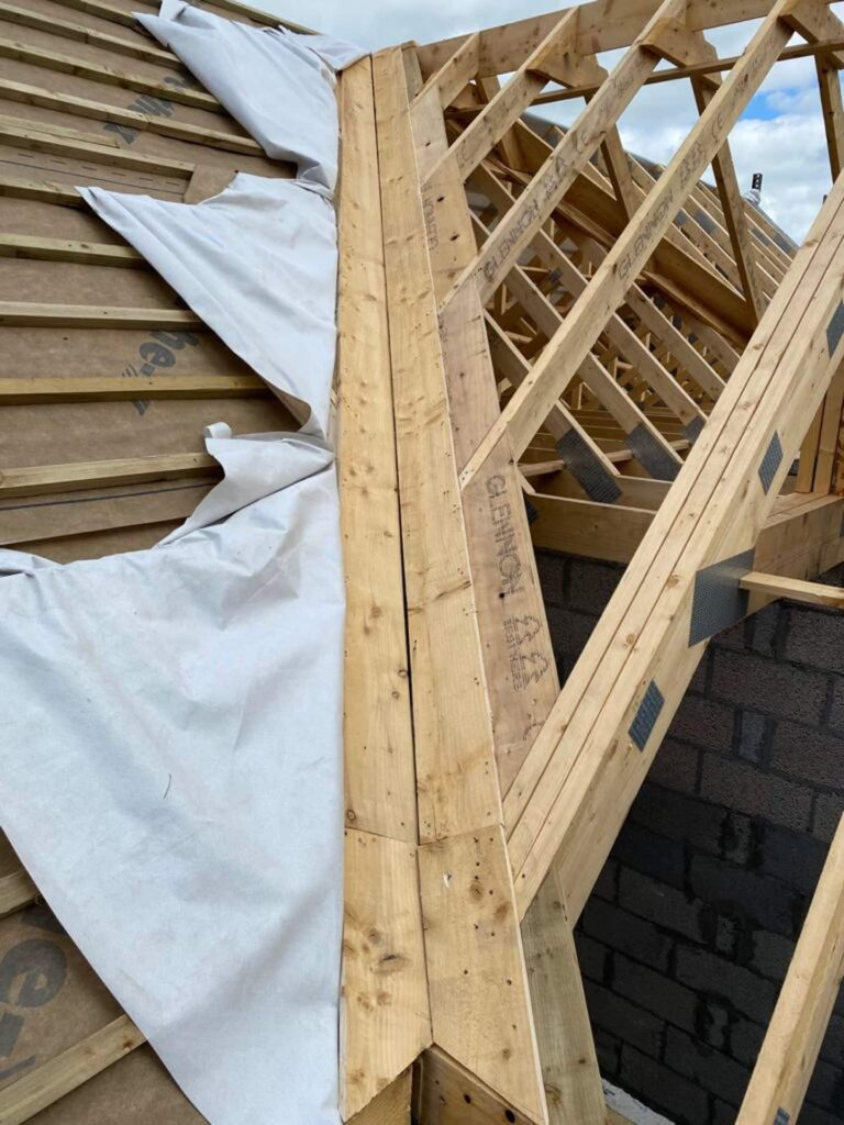 Close-up of timber roof trusses branded Glennon at a hip and valley junction on a new-build residential property in Ireland, with breathable underlay partially unrolled on the adjoining roof section and concrete block gable wall visible below.
