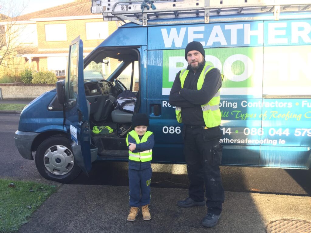 A Weathersafe Roofing tradesperson in a black beanie hat and high-visibility vest standing with arms crossed beside a young child in a miniature high-visibility vest, both posing in front of an early blue Weathersafe Roofing branded van parked on a suburban street in Ireland.
