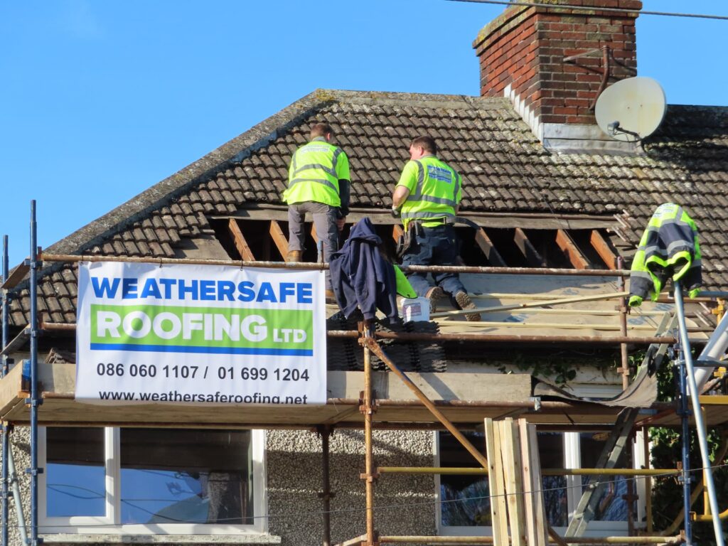 Three Weathersafe Roofing tradespeople in branded high-visibility vests stripping concrete interlocking tiles from the front pitch of a single-storey rendered bungalow in Ireland, with stripped timber battens exposed and the branded Weathersafe Roofing Ltd site board displayed on the scaffold below.