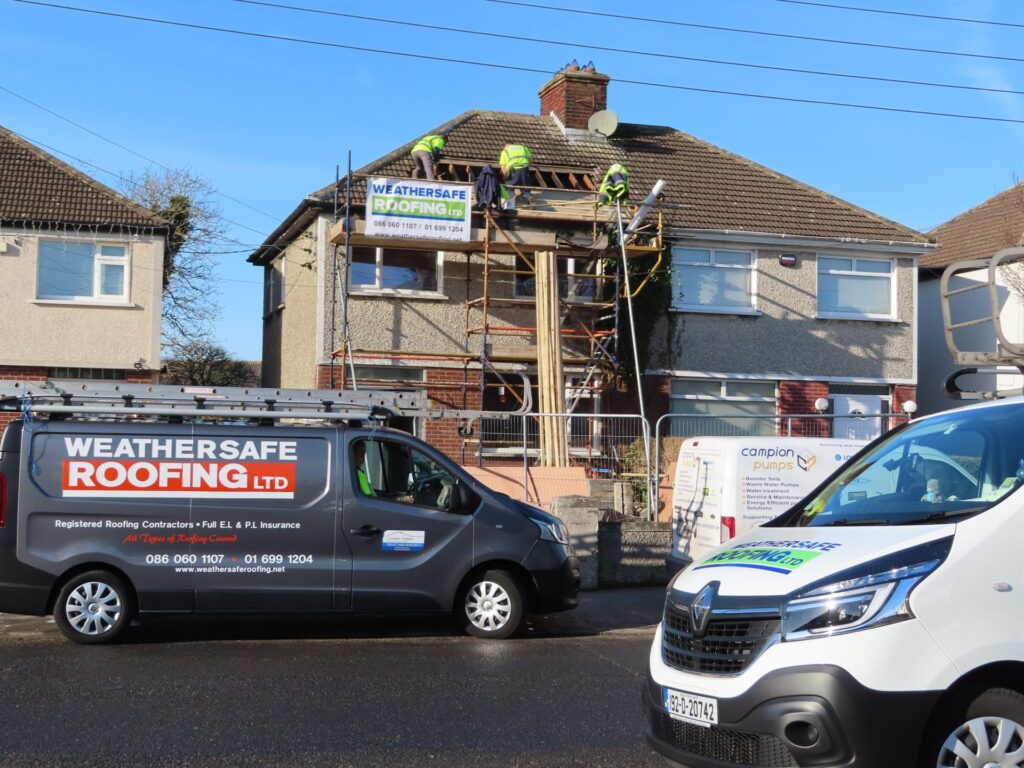 Three Weathersafe Roofing tradespeople in high-visibility vests stripping concrete tiles from the roof of a two-storey semi-detached house in a Dublin suburb, with scaffold erected and the branded Weathersafe Roofing Ltd site board displayed, and two branded company vans parked on the road below.