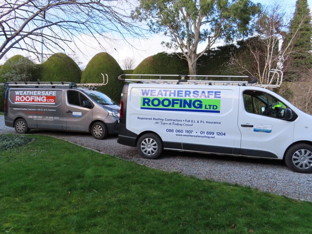 Two branded Weathersafe Roofing Ltd vans — one white and one grey — parked on a gravel driveway at a residential property in Ireland, both displaying the company name, phone numbers 086 060 1107 and 01 699 1204, and website address, with roof racks fitted and garden hedging and trees visible in the background.