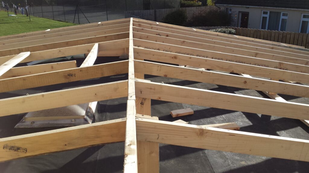 Aerial view of a newly erected CE-graded dry graded timber cut roof frame on a community or commercial building in Ireland, showing ridge board and rafters spanning from the ridge to the eaves over an existing flat roofing membrane, with a sports pitch, fencing and a single-storey outbuilding visible beyond.