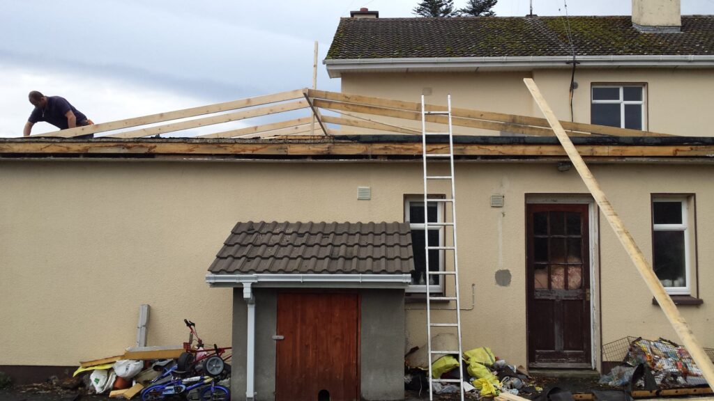 Front elevation of a single-storey rendered residential property in Ireland at early roof replacement stage, with a roofer visible on the roof laying new timber rafters over the stripped flat roof deck, ladders and timber battens staged at ground level, and a concrete interlocked garage roof visible to the centre.