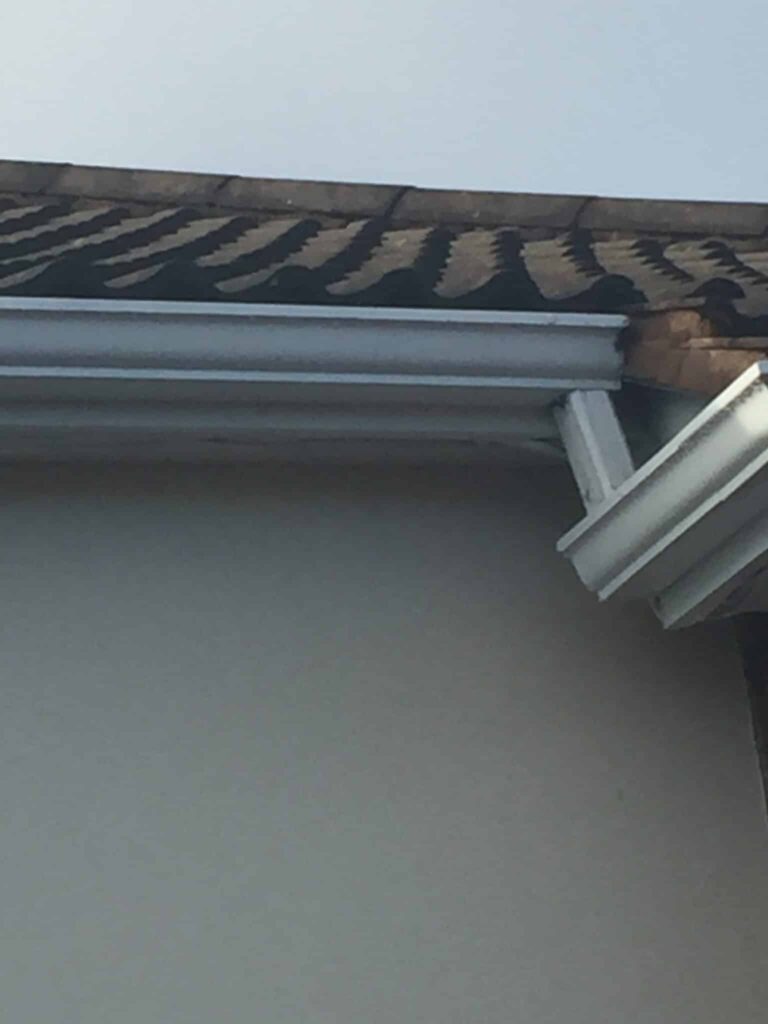 Low-angle view of an eaves corner on a rendered house. A new grey UPVC square-profile gutter with an external corner piece and downpipe is fitted below an aged concrete interlocking tile roof. The gutter outlet and the start of the downpipe run are clearly visible. The rendered wall is visible below.