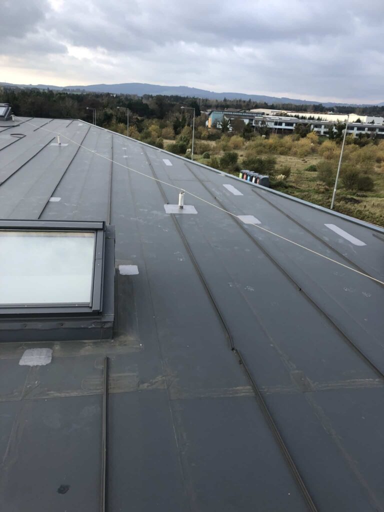 Elevated view of a large commercial flat roof in grey standing seam metal, showing a framed rooflight unit in the lower left corner, a central vent pipe with base flashing, and multiple white seam repair patches. A safety line is visible running diagonally across the roof. Beyond the parapet, scrubland, commercial buildings, and the Dublin mountains are visible under a grey overcast sky.