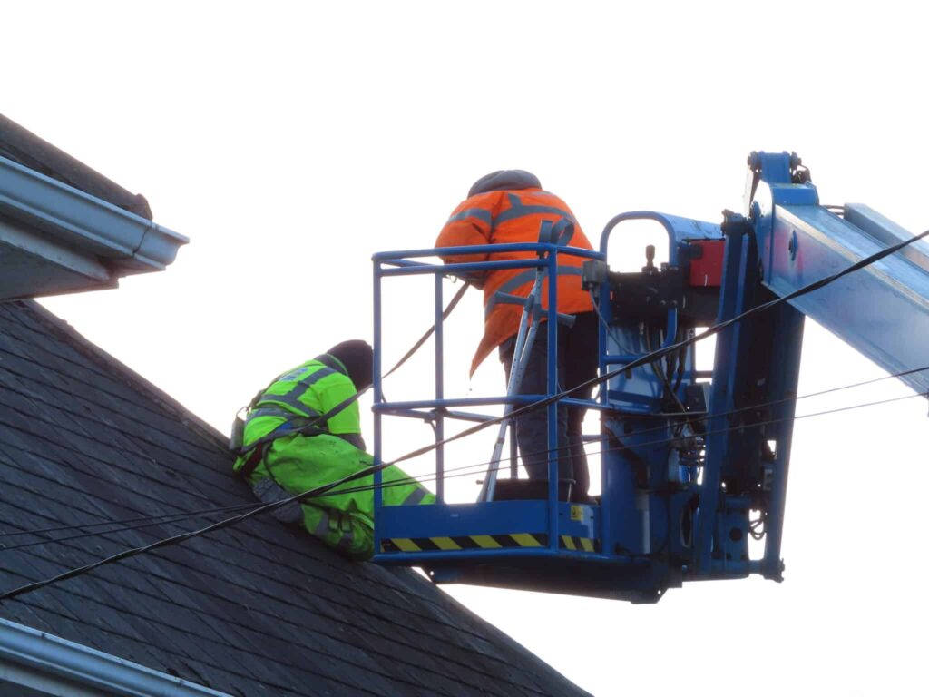 Two roofers working at roof ridge level. One worker in a lime green high-visibility jacket crouches on the slate roof slope; another in an orange high-visibility jacket stands in the platform basket of a blue cherry picker elevated work platform positioned at the ridge. Both are working on the same area of the roof against a bright white overcast sky.