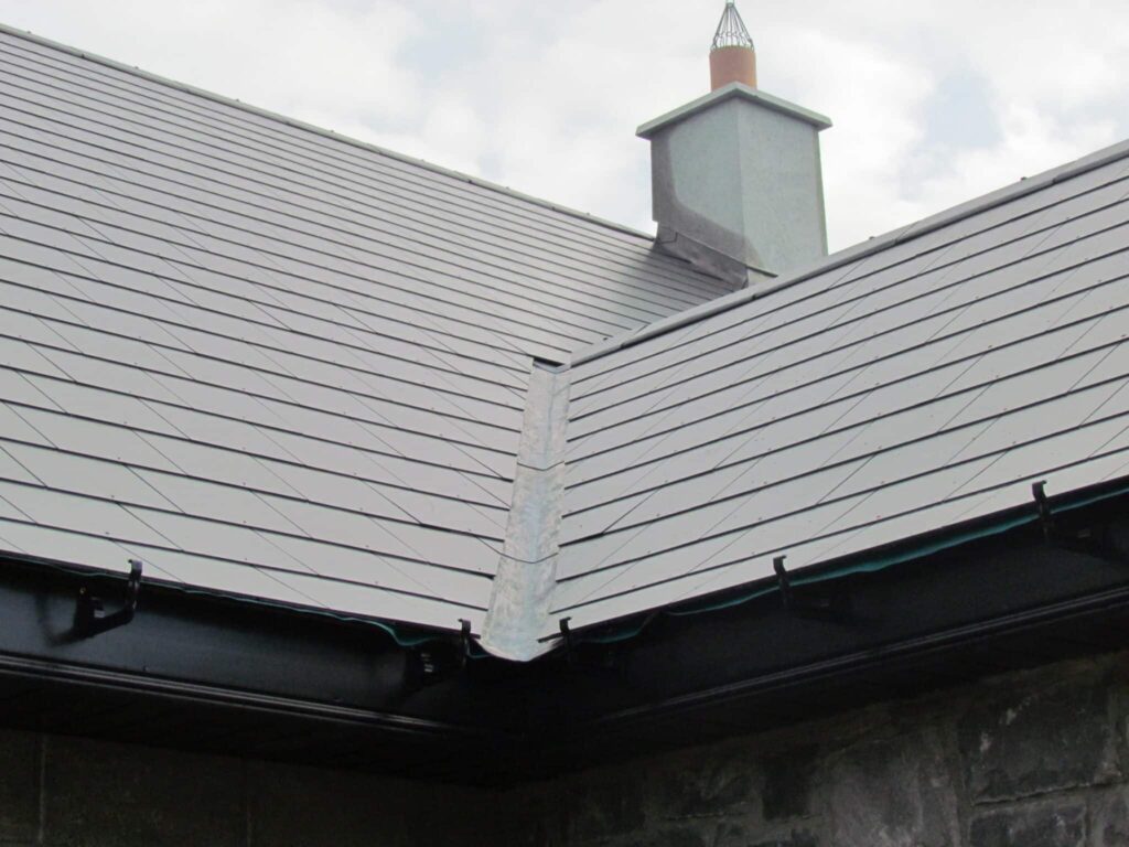 Exterior view of a completed natural slate roof on a single-storey building, showing a hipped corner detail with a neat lead or zinc hip flashing strip running down the junction of the two slated pitches. Black cast-iron style guttering runs along the eaves on both visible sides. A rendered and painted chimney stack with a bird-guard chimney pot is visible above against an overcast sky.