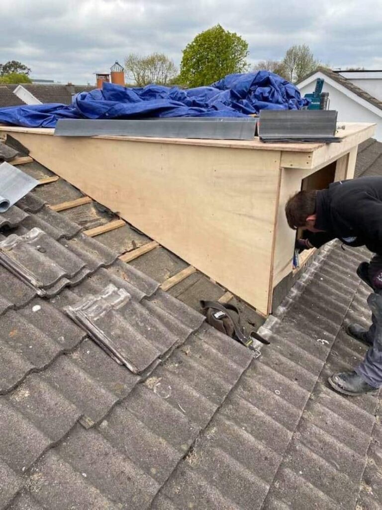 A roofer crouches at the side opening of a plywood flat-roof dormer box, working on the internal junction. Cut sheets of zinc or lead cladding material are stacked on top of the dormer ready for fixing. The existing concrete tile roof is visible in the foreground and on the far side of the dormer. A blue tarpaulin partially covers the dormer top.
