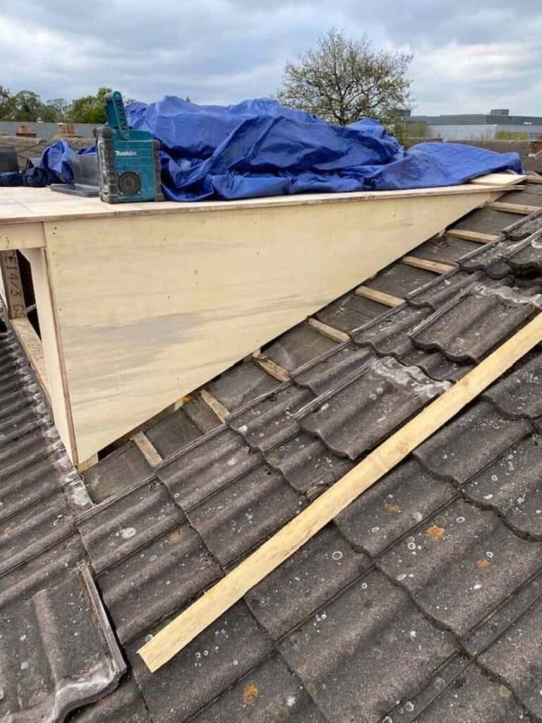 Side-on view of a newly built flat-roof dormer box clad in plywood sheeting on a concrete tile roof slope. A blue tarpaulin and Makita power tool are visible on top of the structure. Timber battens and part of the existing tile roof are visible in the lower foreground. A suburban skyline with trees and buildings is visible in the background.