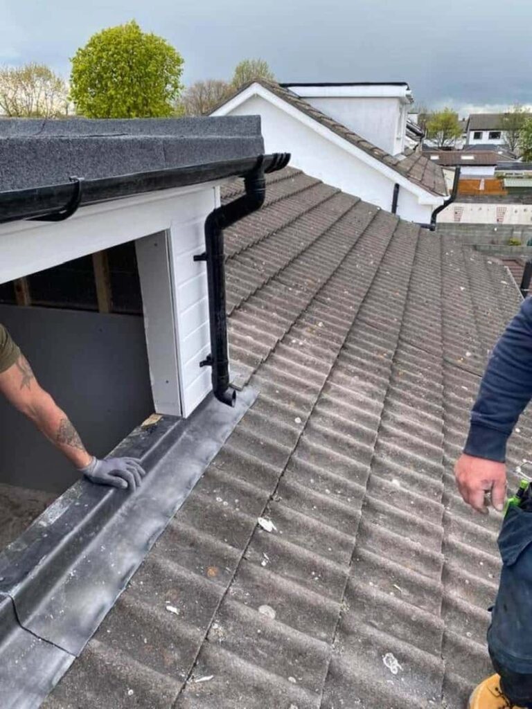 Two roofers work together on a concrete interlocking tile roof beside a white-rendered dormer structure. One roofer, wearing gloves, crouches to dress and fix a lead or zinc apron flashing strip at the base of the dormer abutment. A black UPVC downpipe and guttering are visible on the dormer. Neighbouring rooftops and trees are visible in the background under a cloudy sky.