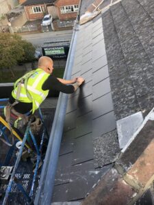 A Weathersafe Roofing worker in a yellow high-visibility vest stands on a blue scaffold platform, leaning forward to carefully lay and align dark grey synthetic slate tiles at the eaves of a roof. A grey aluminium gutter runs below. The aged existing slates are visible alongside the new section. A Weathersafe Roofing Ltd sign is visible on a van parked in the street below. A residential street, parked cars, and a brick building are visible in the background.
