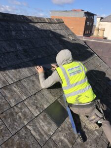 A roofer wearing a yellow Weathersafe Roofing Ltd high-visibility vest and grey hoodie crouches on an aged, lichen-covered slate roof, carefully fitting a new replacement slate into a gap. A second new slate is staged nearby. A brick building and chimneys are visible in the background under a blue sky.