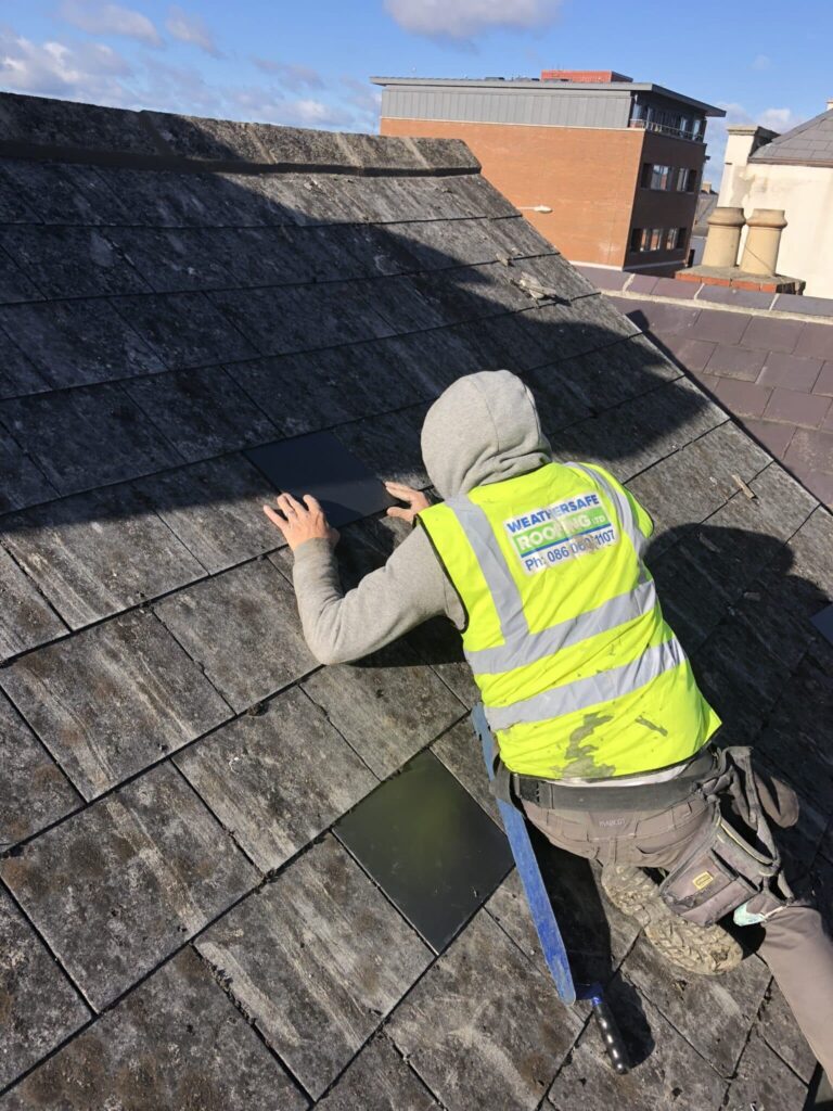 A roofer wearing a yellow Weathersafe Roofing Ltd high-visibility vest and grey hoodie crouches on an aged, lichen-covered slate roof, carefully fitting a new replacement slate into a gap. A second new slate is staged nearby. A brick building and chimneys are visible in the background under a blue sky.