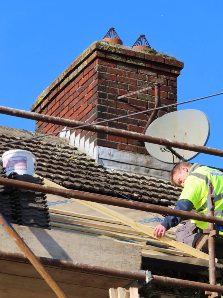 A roofer wearing a yellow high-visibility vest kneels on scaffolding boards, handling timber roof battens with new felt underlay beneath. A weathered brick chimney stack with bird guard chimney pots and a satellite dish dominates the background against a blue sky.