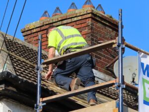 A roofer in a yellow high-visibility vest with the Weathersafe Roofing logo visible on the back, crouching on scaffolding boards beside a large brick chimney stack. The tiled roof and chimney pots with bird guards are visible above against a clear blue sky.