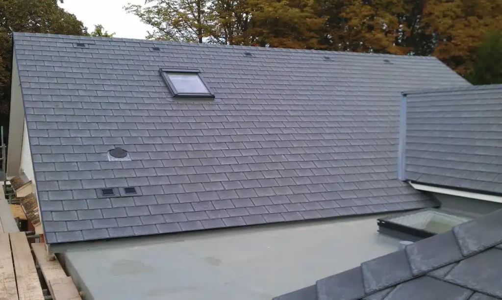 Elevated view of a completed residential roof showing a large pitched section covered in mid-grey slate tiles with a white PVC rooflight window, two slate vent tiles, and eaves ventilation grilles. An adjacent flat roof section with grey membrane and a small rooflight is visible in the lower right. Autumn trees are visible in the background.