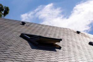 Low-angle view looking up a grey natural slate pitched roof. A black-framed rooflight window is hinged open. Two roofline vent terminals are visible further up the slope against a partly cloudy blue sky.