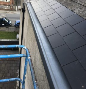 Close-up of a partially reslated roof from scaffolding height. A section of new dark grey/black slate tiles has been laid neatly at the lower eaves, contrasting with the older weathered slates above. A new grey aluminium gutter has been fitted below. A roofer is visible at the top of the image.