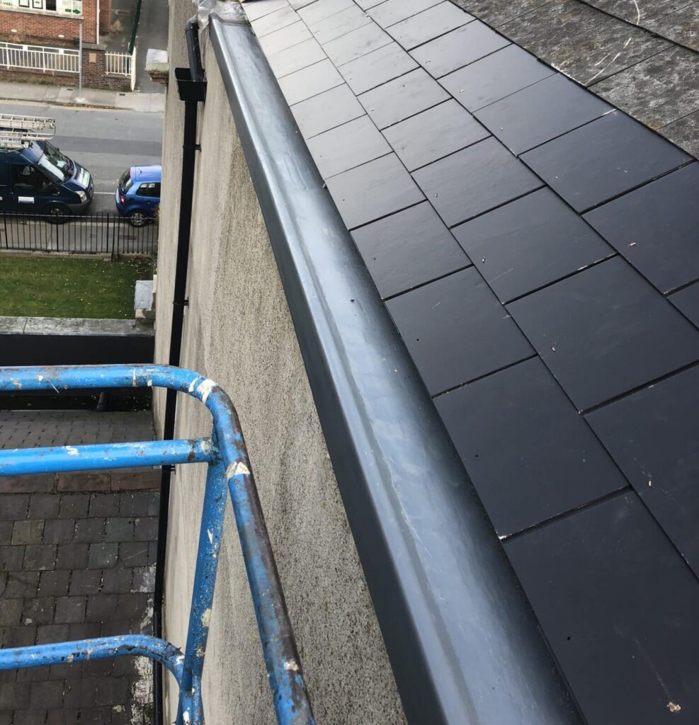 Close-up of a partially reslated roof from scaffolding height. A section of new dark grey/black slate tiles has been laid neatly at the lower eaves, contrasting with the older weathered slates above. A new grey aluminium gutter has been fitted below. A roofer is visible at the top of the image.