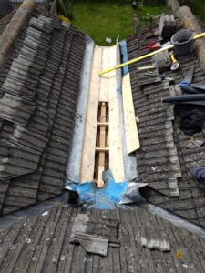 Overhead view of a roof valley mid-repair. Old concrete tiles have been stripped back on both sides, exposing timber joists and new boarding in the valley. Blue and silver flashing materials are visible. Tools and a bucket are present on the roof surface.