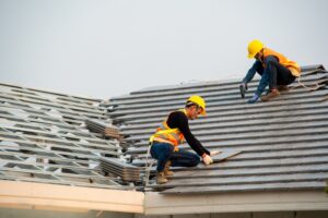 Two roofers in yellow hard hats and high-visibility vests work together on a steeply pitched roof, laying and securing grey concrete roof tiles. Stacked tiles are visible in the foreground against a pale sky.