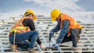 Two roofers in high-visibility orange vests and yellow hard hats work on a metal roof frame structure. One uses a power drill while kneeling on metal joists against a partly cloudy sky.