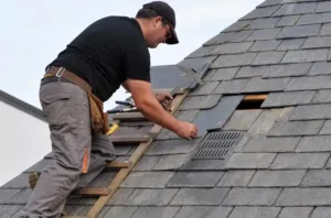 A roofer in a black cap and work trousers, standing on a wooden roof ladder, lifts and repositions a slate tile on a grey slate pitched roof. A vent tile is visible nearby.