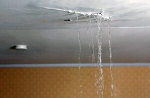 Close-up of a ceiling with an active roof leak. Multiple streams of water pour down through a hole in a grey suspended ceiling panel, with visible pooling above.